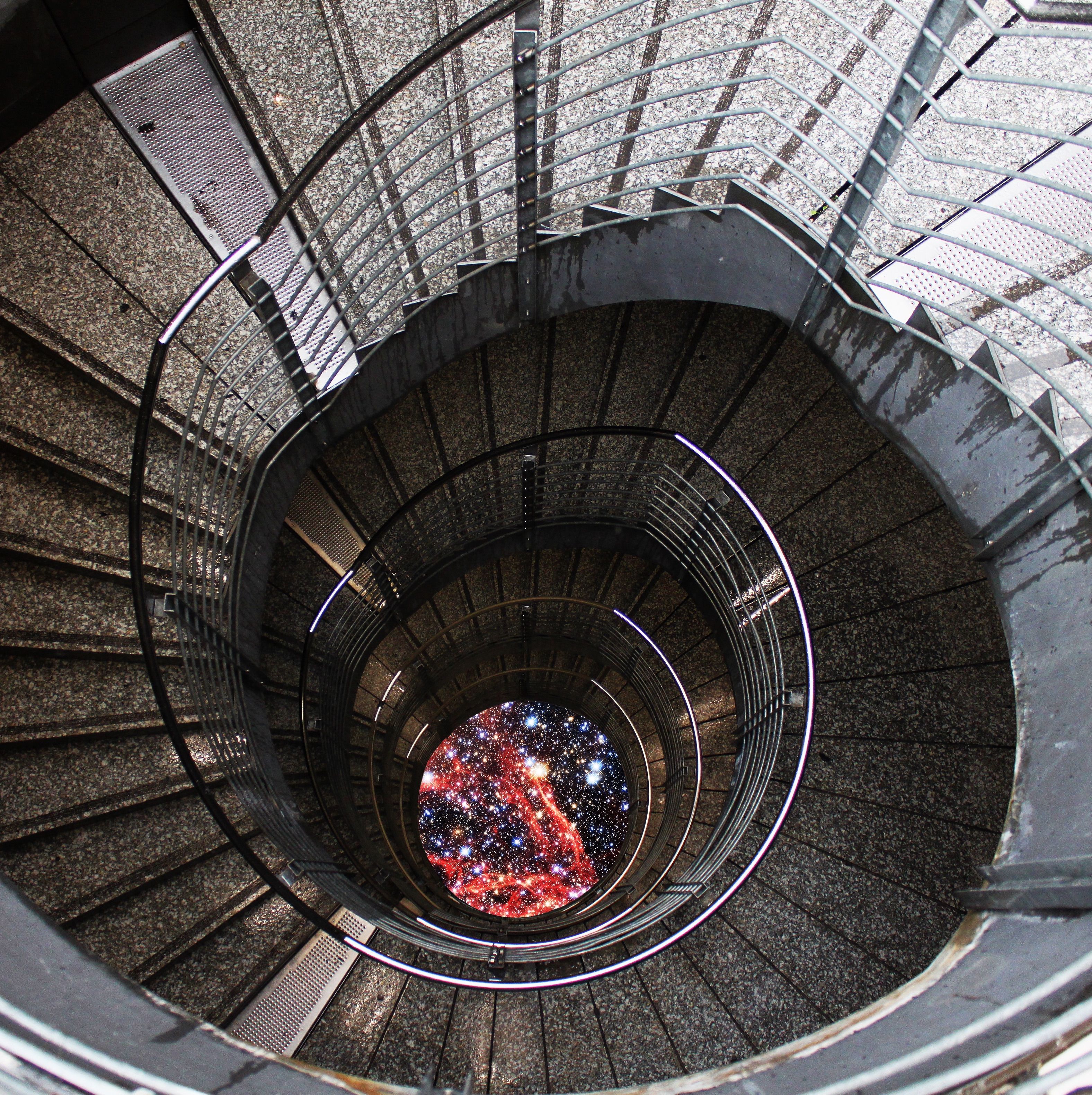 Aus der Vogelperspektive sieht eine:r eine Wendeltreppe, die nach unten führt. Anstelle des Bodens sieht eine:r ein Bild vom Universum in der Mitte der Treppe.