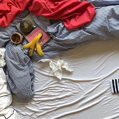 Ein Bett aus einer Vogelperspektive. Darauf blau gestreifte zerknautschte Bettwäsche, ein Kaffeebecher, eine Bananenschale und ein rotes Tuch.