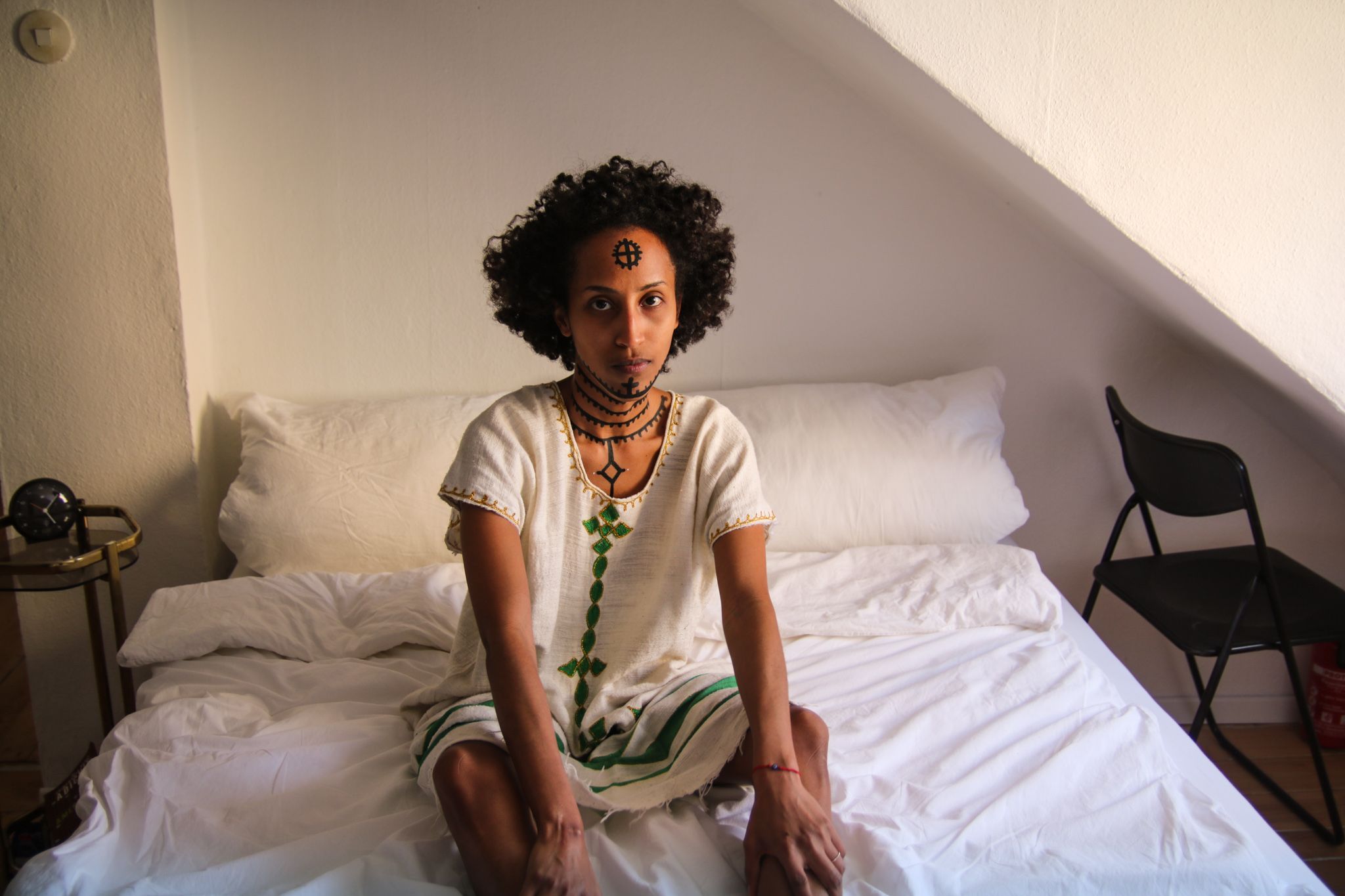 A woman is sitting on the edge of a white covered bed. Her upper body and head are turned sideways to the left behind her. She is looking up, directly into the camera. Her neck, chin and forehead are painted with traditional Ethiopian tattoos.