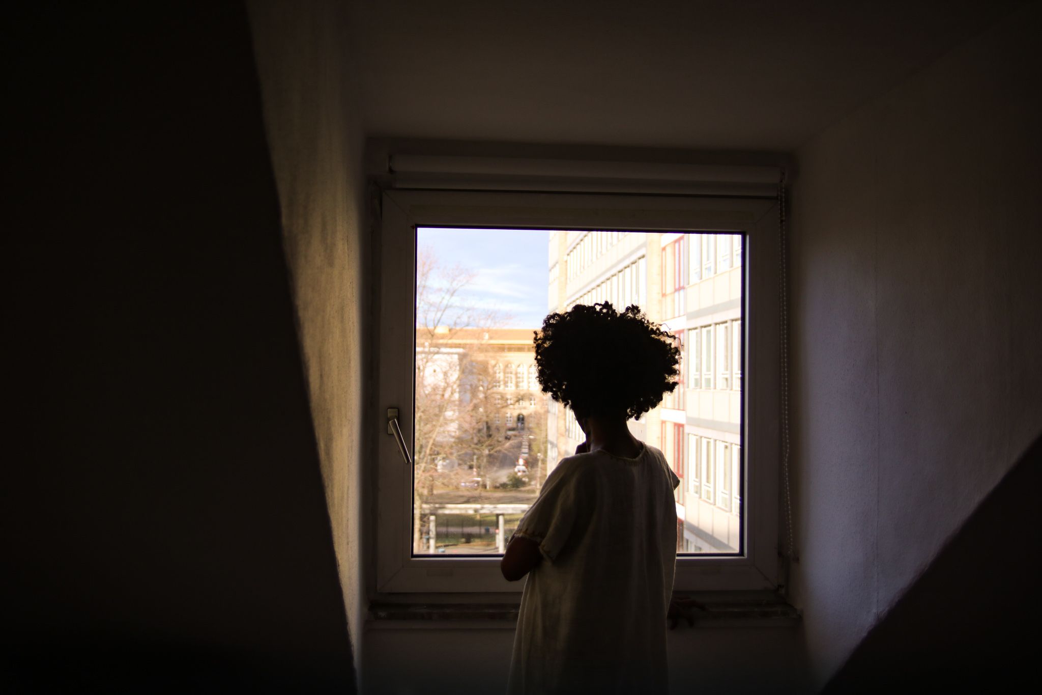 Die obere Hälfte einer Person ist stehend vor einem Fenster zu sehen. Es ist ihr weiß gekleideter Rücken zu sehen. Die Person ist nur schemenhaft zu erkennen. Rechts und Links von ihr sind Dachschrägen. Es fällt wenig Licht ein.