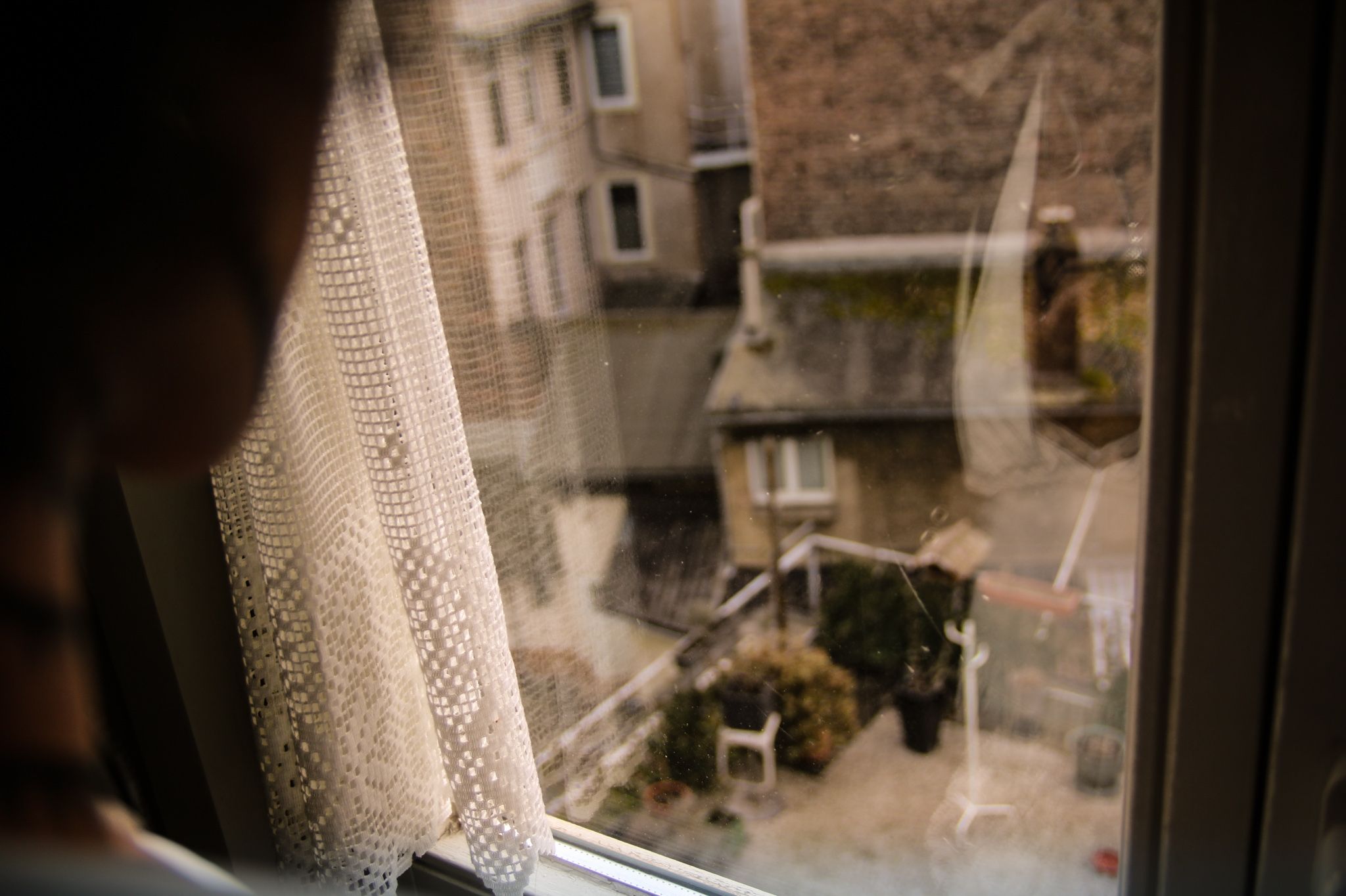 Links im Bild ist verschwommen der Rand von einem Gesicht, Hals und einer Schulter. Man blickt aus einem Fenster in einen kleinen Hof mit Terrasse, Pflanzen, Dächern und Häuserwänden. Eine Gardine ist am Fenster, aus dem man blickt, zur Seite gezogen.