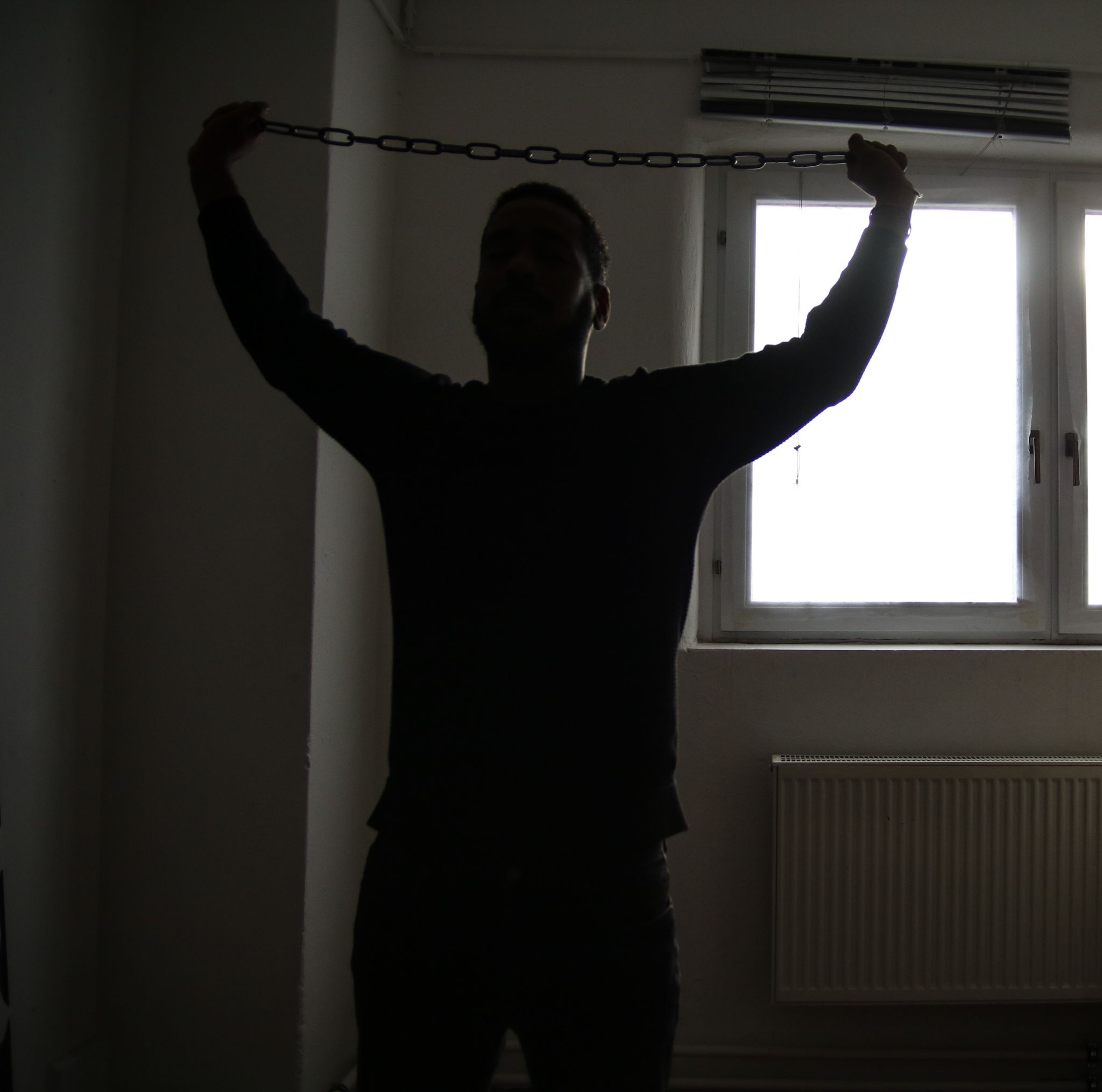 Im Mittelpunkt steht die frontale Silhouette eines Mannes. Schwaches Licht durch ein Fenster im Raum. Über seinem Kopf hält er zwischen seinen erhobenen Armen eine gespannte Metallkette.