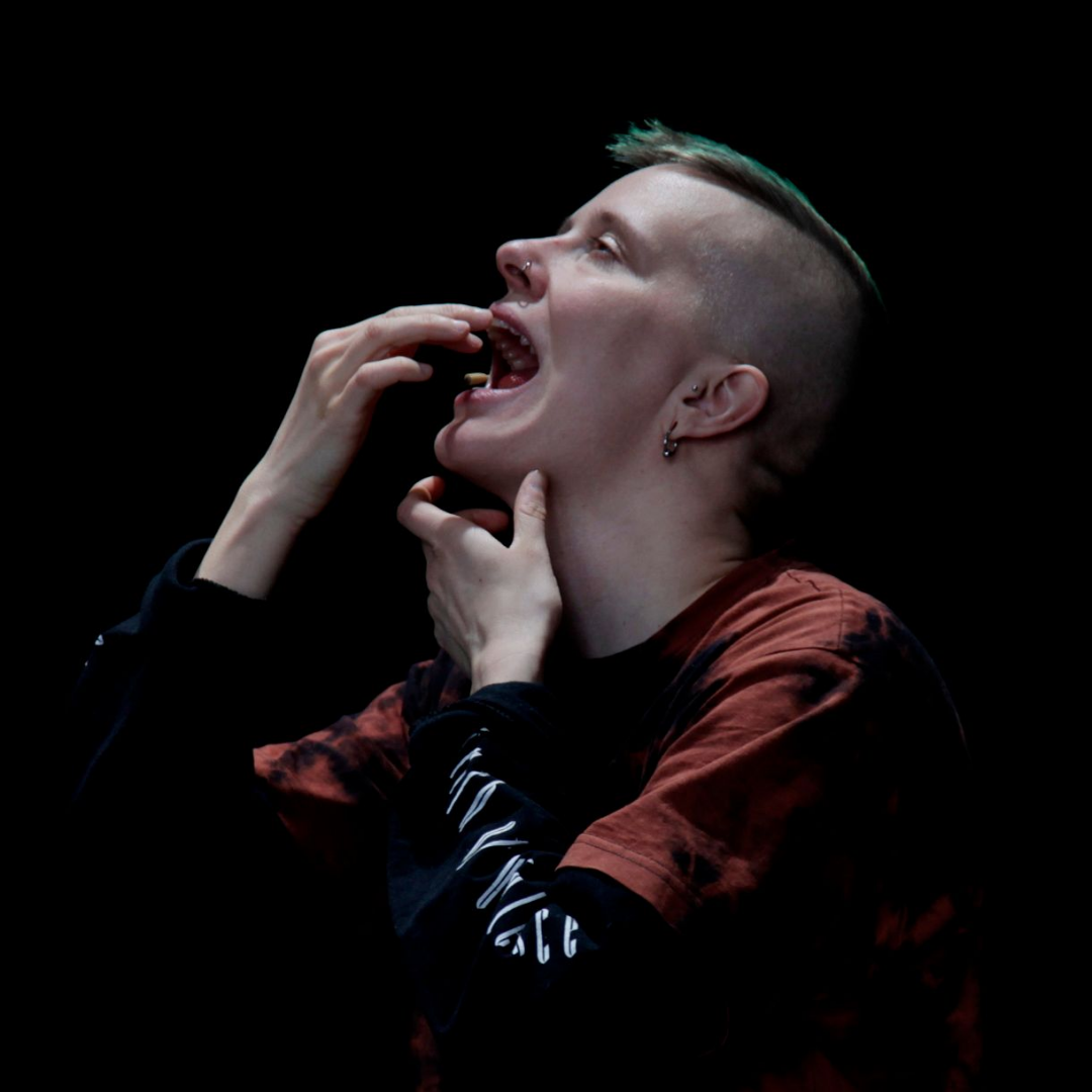 Vor einem schwarzen Hintergrund sieht eine:r Performer:in Teo Ala-Ruona von der Seite. Ala-Ruona fasst sich mit der linken Hand an die Kehle. Die rechte Hand ist vor dem geöffneten Mund.