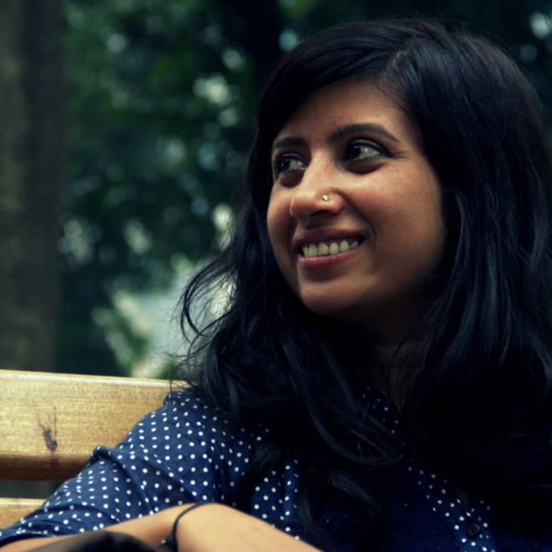 Neha Singh sitzt auf einer Bank und hat ihren Blick nach links oben gerichtet. Im Hintergrund sind grüne Baumkronen zu sehen.
